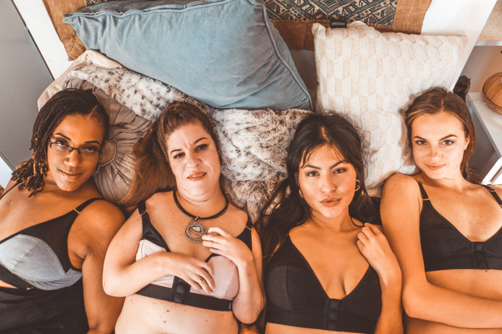 Photo of 4 women wearing Elba London bras, lying down, looking up at the camera on a bed with a number of colourful cushions.